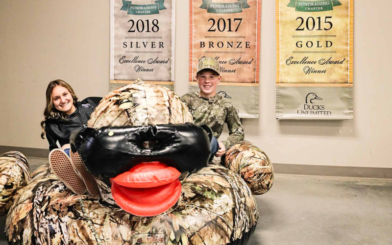 Mariah Messmer and Sutten Jensen pose for a photo atop an inflatable duck donning shades, one of the many Ducks Unlimited auction items. Banners touting national recognition for the local chapter are displayed behind the duo. The chapter recently received the top honor of “Elite” status for its work during 2021.