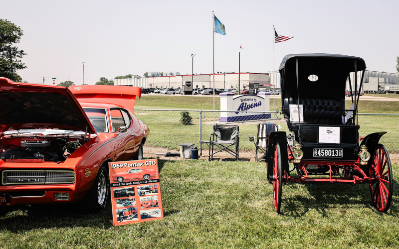 The Alpena Town sign is perched between two of the car show entries, a 1909 Sears Motor Buggy (right) and a 1969 Pontiac GTO.