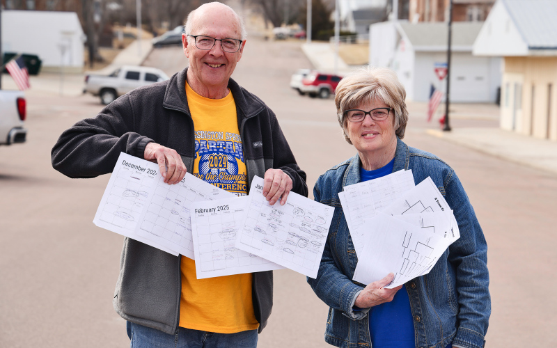 KRISTI HINE / TRUE DAKOTAN Mike and Terri Mebius hold stacks of detailed sports calendars and basketball tournament brackets as they stand on Main Street in Wessington Springs. They use the tools to guide them across the state as they follow their grandchildren play basketball. In an exciting turn of events, all three of their grandsons’ varsity teams made it to the state tournaments, scheduled to be held in Aberdeen, Sioux Falls and Rapid City.