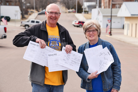 KRISTI HINE / TRUE DAKOTAN Mike and Terri Mebius hold stacks of detailed sports calendars and basketball tournament brackets as they stand on Main Street in Wessington Springs. They use the tools to guide them across the state as they follow their grandchildren play basketball. In an exciting turn of events, all three of their grandsons’ varsity teams made it to the state tournaments, scheduled to be held in Aberdeen, Sioux Falls and Rapid City.